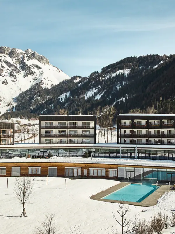 Badewelt im aja Bergresort Werfenweng Das Hotelgebäude mit dem Außenpool im Schnee, umgeben von Bergen.