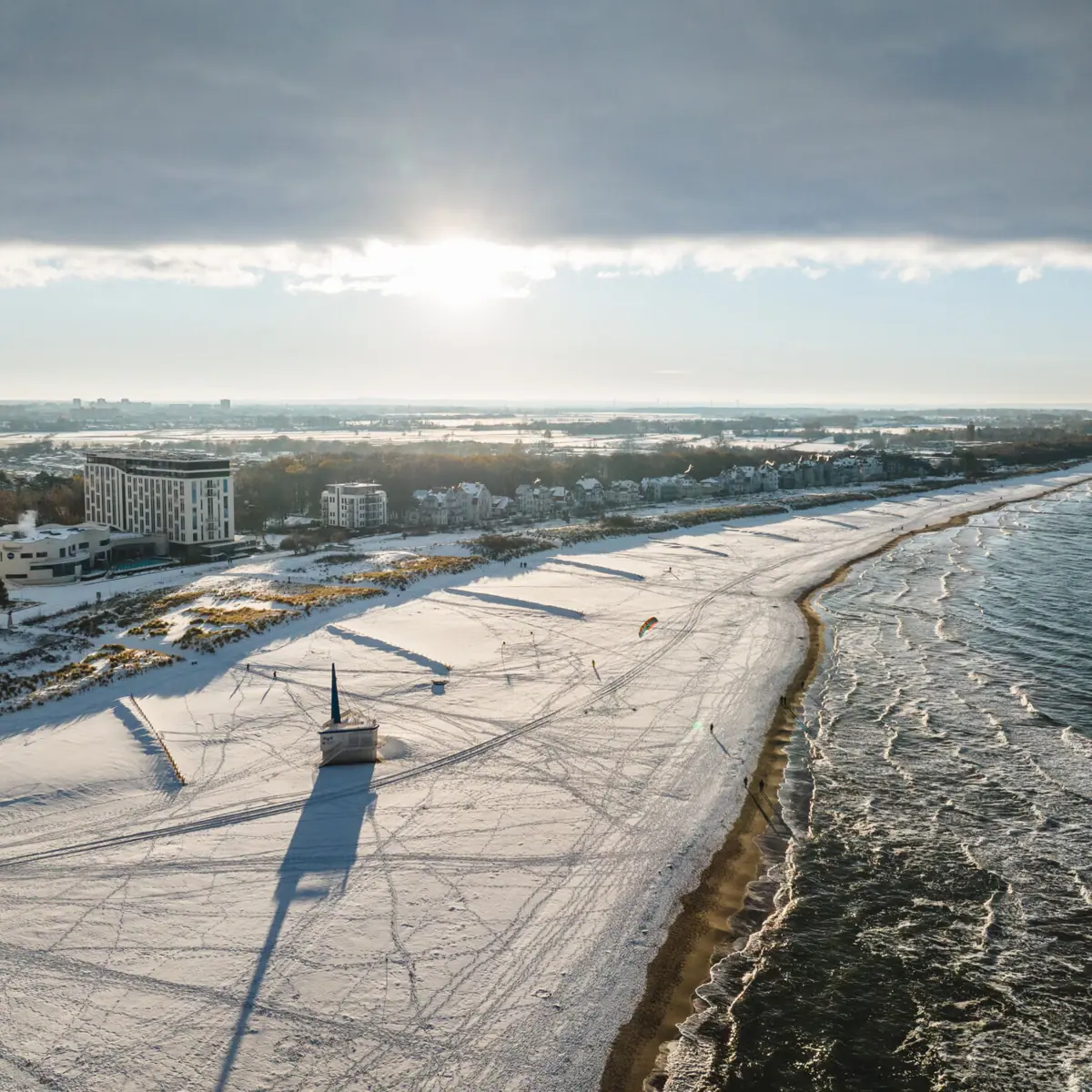 Ein schneebedeckter Strand in Warnemünde im Winter mit Gebäuden und Wasser im Hintergrund.