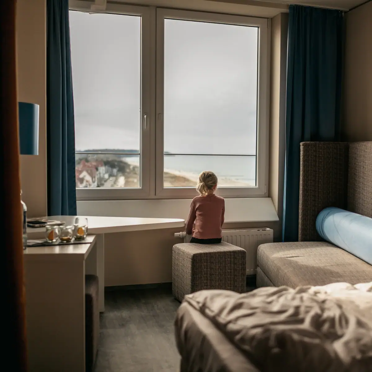 Zimmer im aja Warnemünde Ein Mädchen sitzt auf einem Hocker und schaut aus dem Fenster.