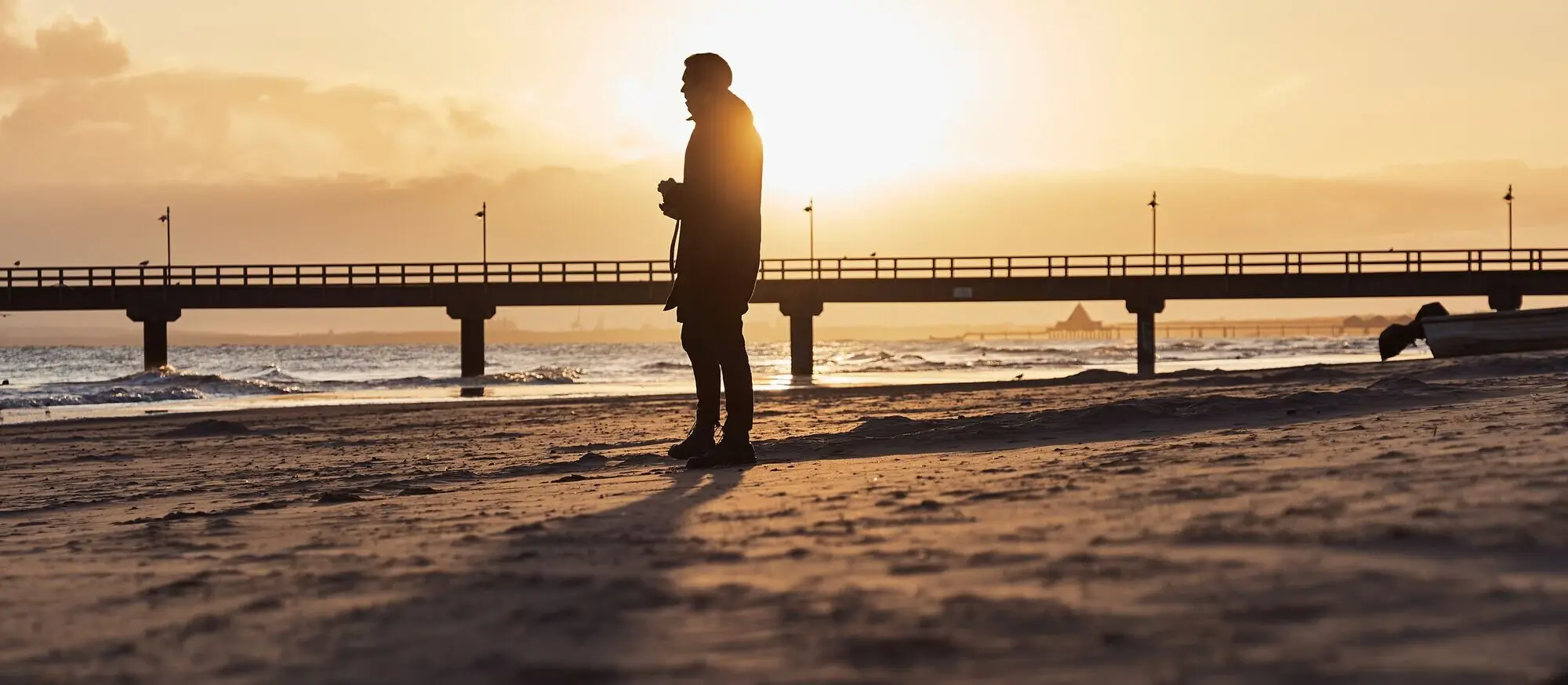 Man standing on a beach with a bridge in the background.