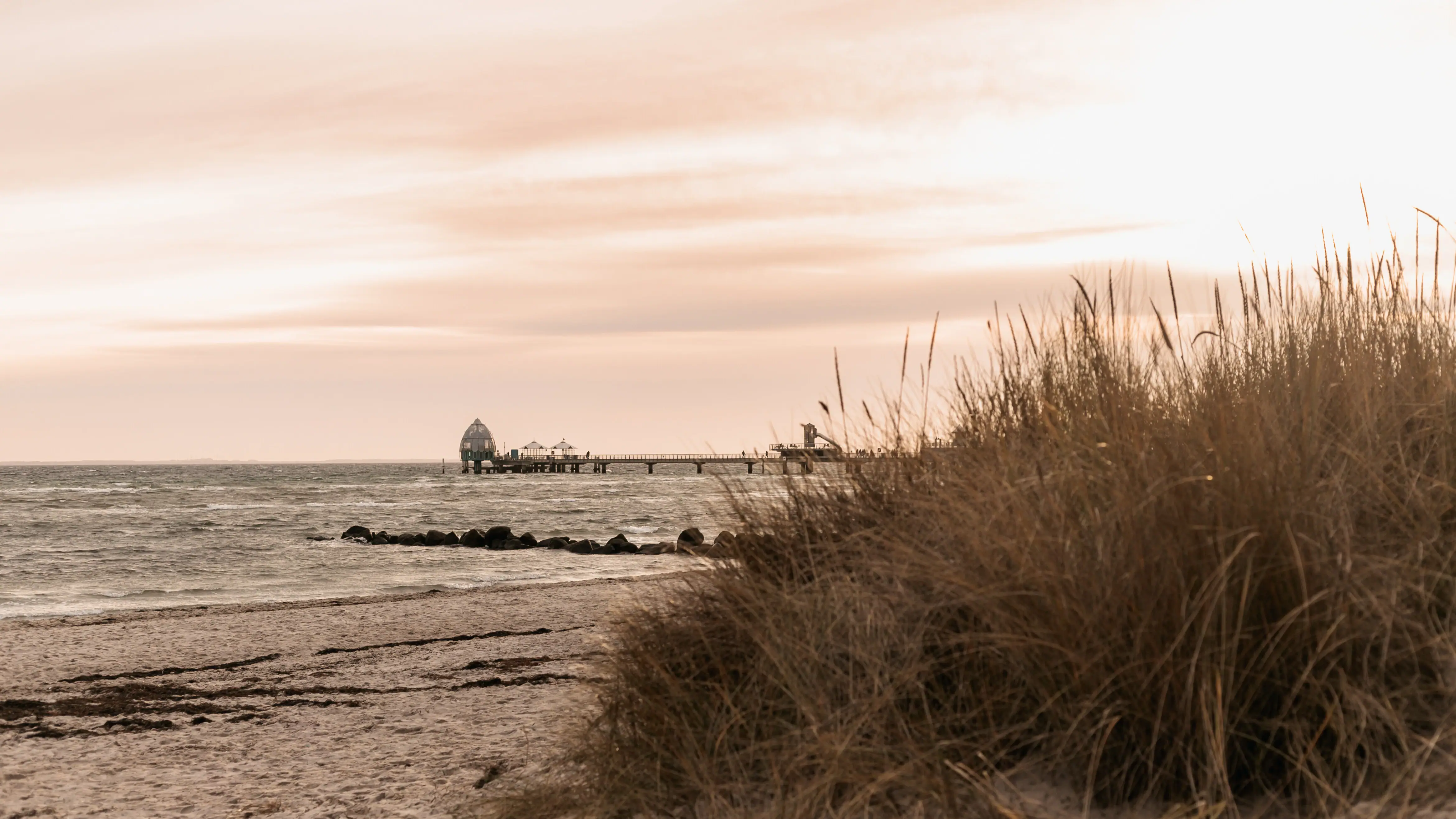 Strand mit einem Pier und Gras im Vordergrund.