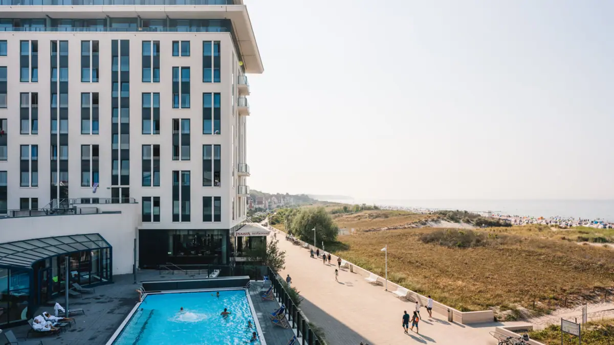 aja Warnemünde Außenansicht mit dem Pool im Vordergrund sowie der Promenade und dem Strand auf der rechten Seite.