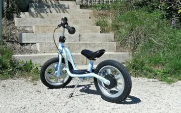 Ein kleines hellblaues Kinderlaufrad steht auf einem Kieselweg vor einer Steintreppe in der Sonne.