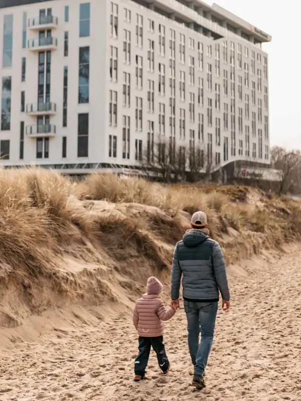 Strandaufgang Eine Frau und ein Kind gehen auf einem unbefestigten Weg mit einem Gebäude im Hintergrund.