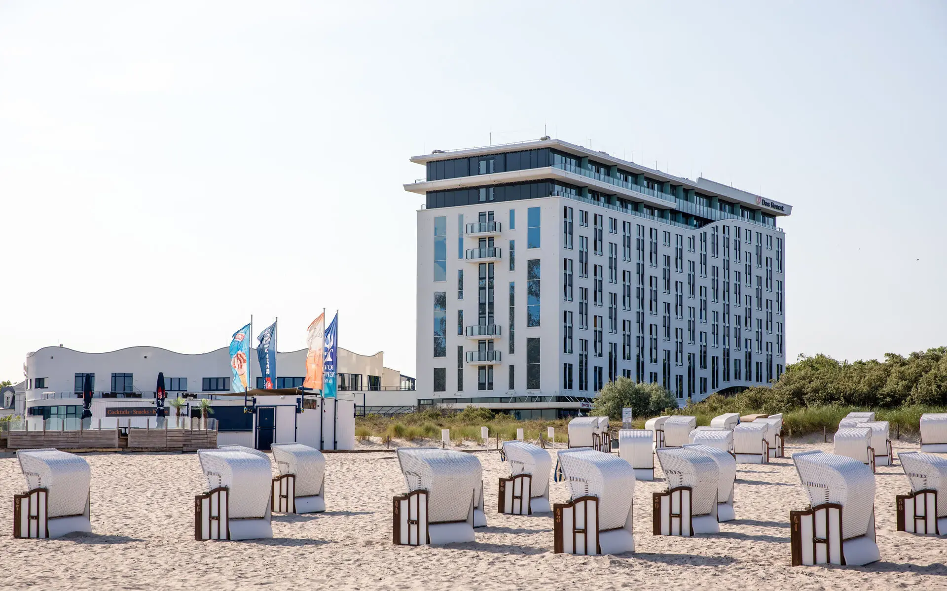 Außenansicht des aja Warnemünde. Auf dem davor liegenden Strand stehen viele Strandkörbe und die Sonne scheint angenehm. 