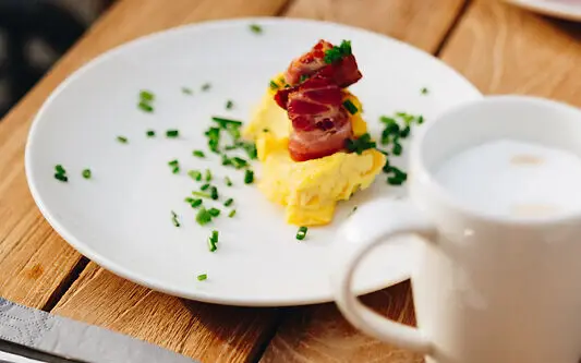 Ein Teller mit Rührei und Bacon auf einem Tisch, umgeben von Besteck und einer Tasse Kaffee.