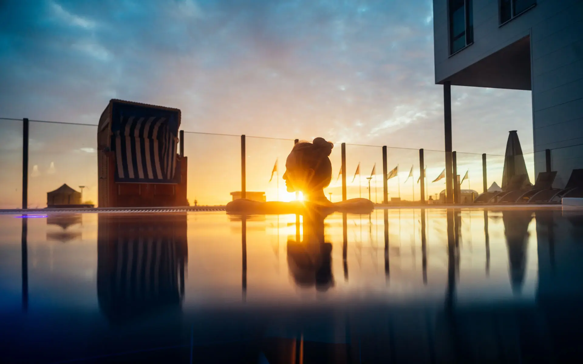 Ein Frau ist bei Sonnenuntergang auf den Beckenrand eines Außenpools gelehnt und schaut in die Ferne. Der Pool ist mit einem Glaszaun begrenzt, dahinter befindet sich das Meer. Davor steht ein blau weiß gestreifter Strandkorb. 