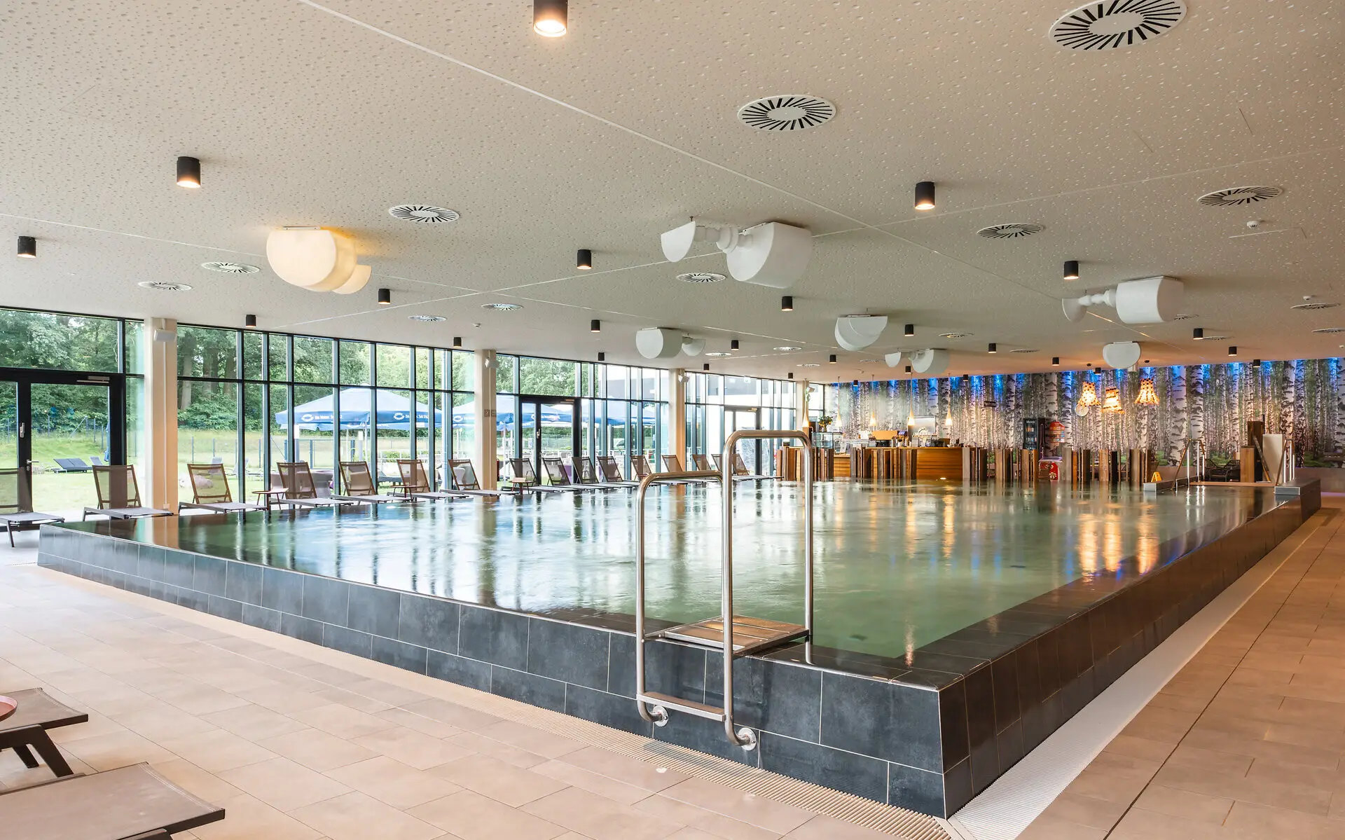 Large indoor swimming pool with a prominent railing and surrounding chairs.