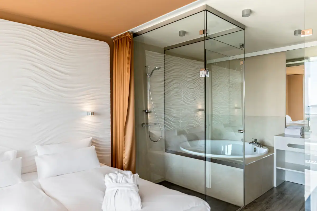 Badezimmer in der Suite im aja Warnemünde Badezimmer mit Glasdusche und Bett im Vordergrund.