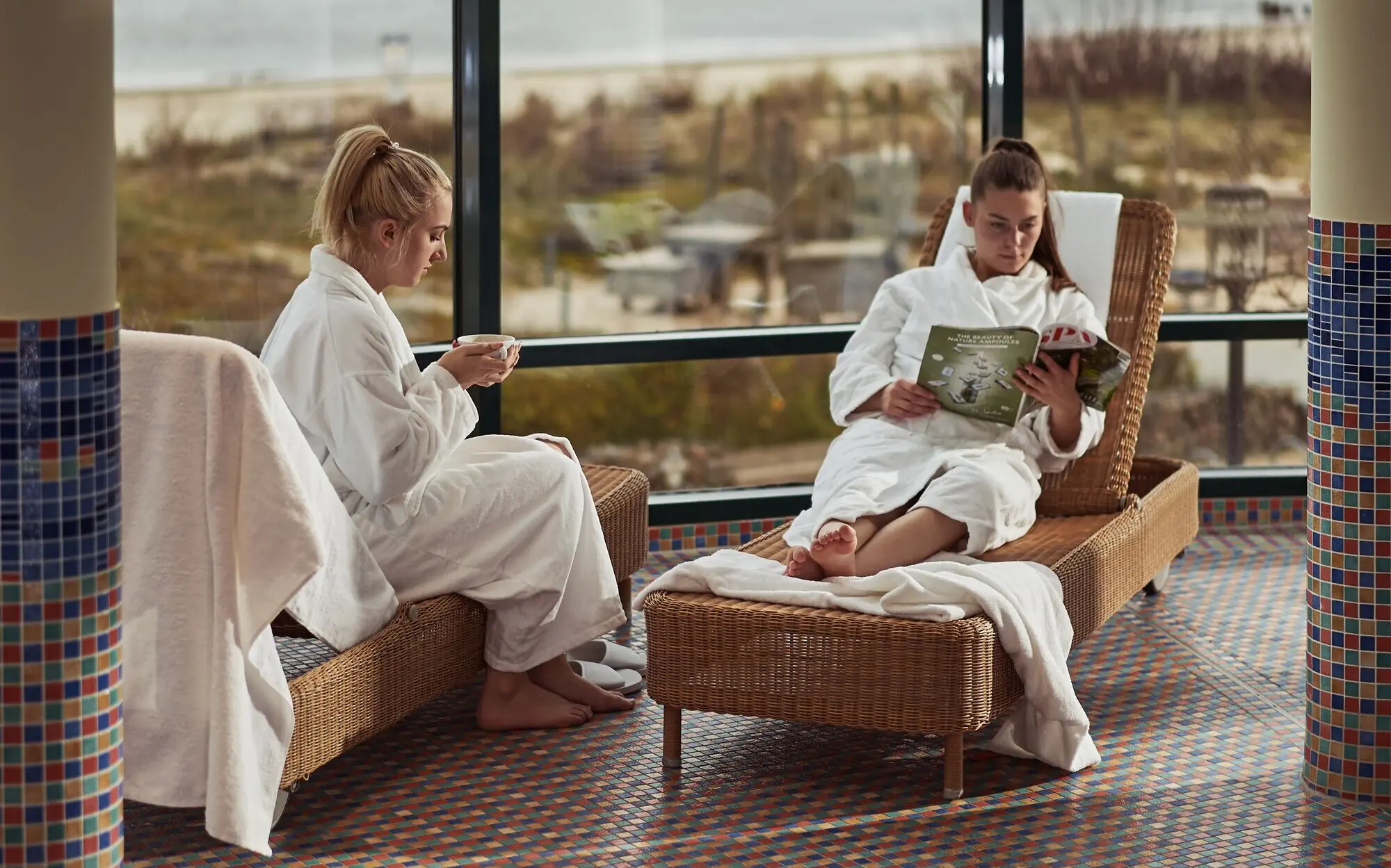 Two women are relaxing in a spa on two loungers overlooking the sea, one holding a cup and the other reading a magazine.