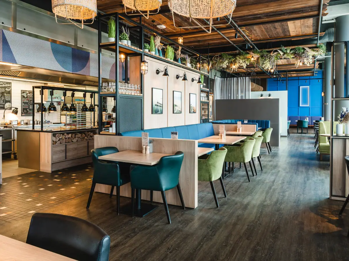 Restaurant im aja Warnemünde Ein Restaurant mit Tischen und Stühlen. Stilvoll und modern eingerichtet. Maritimer Look mit Bildern aus Warnemünde an der Wand. Offene Buffetfläche im Hintergrund.