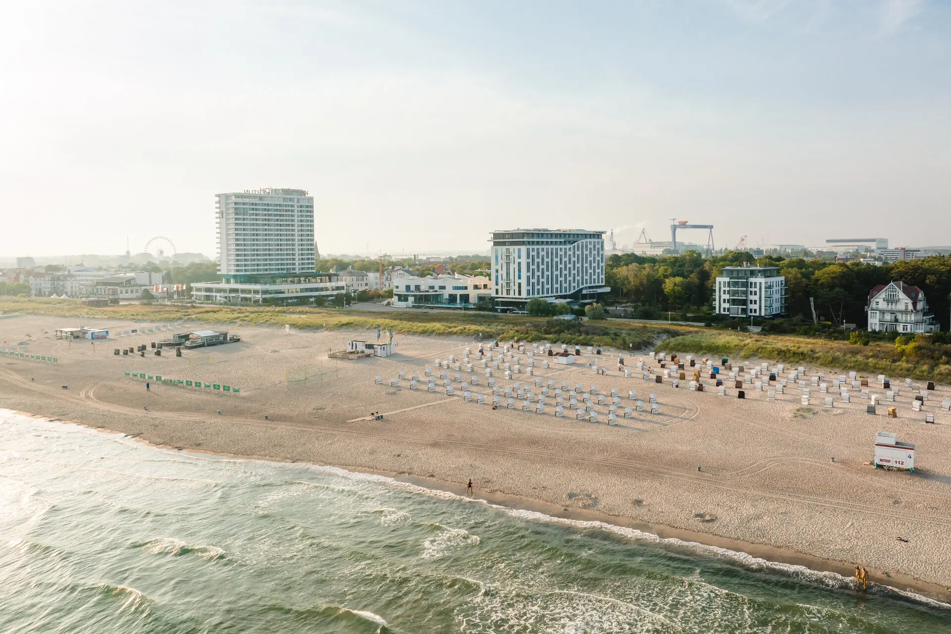 Eine Luftaufnahme eines Strandabschnitts in Warnemünde. Das Meer trifft ruhig auf den breiten Strand mit vielen Strandkörben und Gästen. Dahinter reihen sich verschiedene Gebäude aneinander. Die Sonne scheint warm vom Himmel.