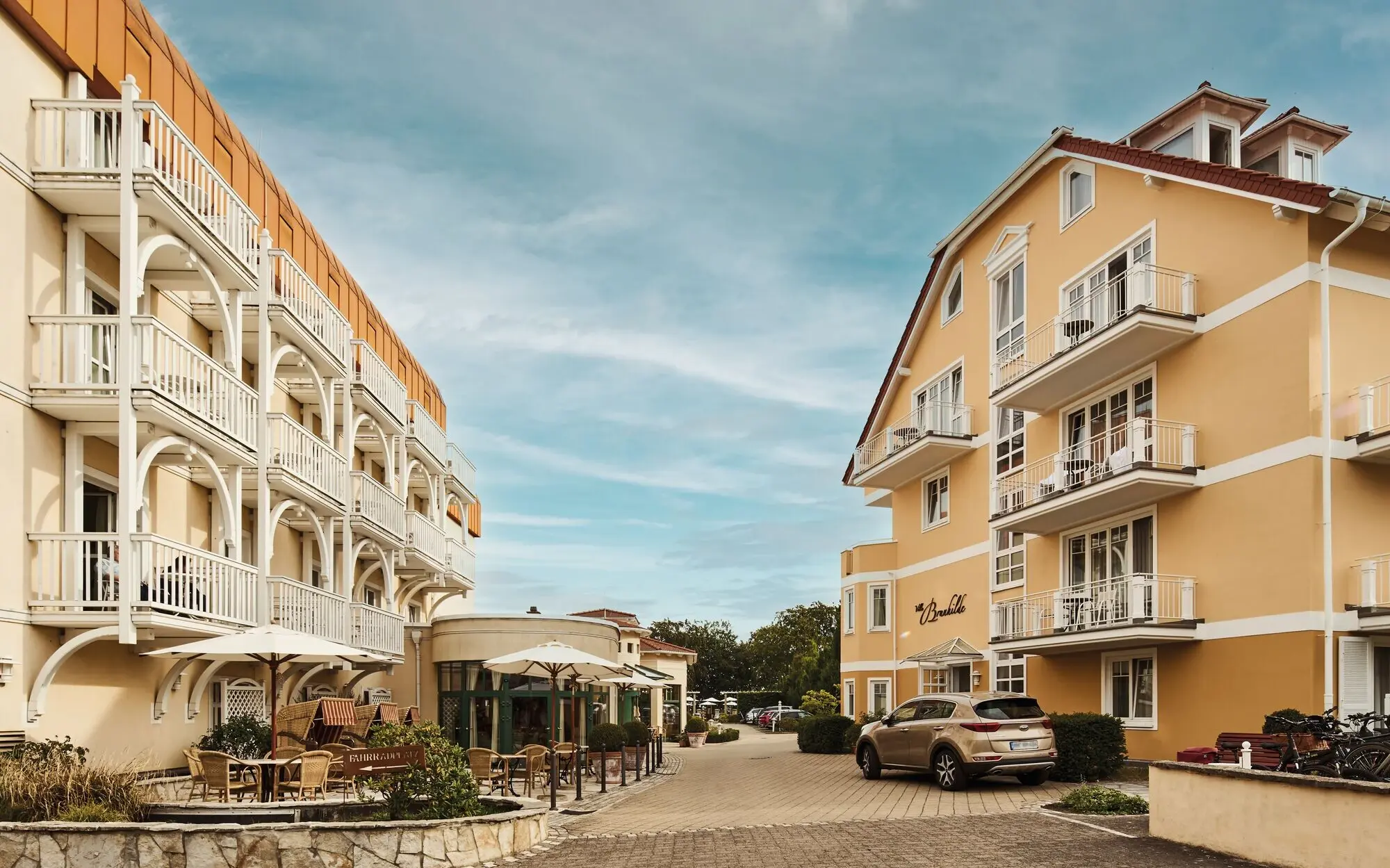 Außenansicht des Nordperd & Villen Göhren mit zwei charmanten Gebäuden im Bäderstil, Balkonen, Sonnenterrasse und einladender Einfahrt; davor geparkte Autos und Sitzgelegenheiten unter Sonnenschirmen.