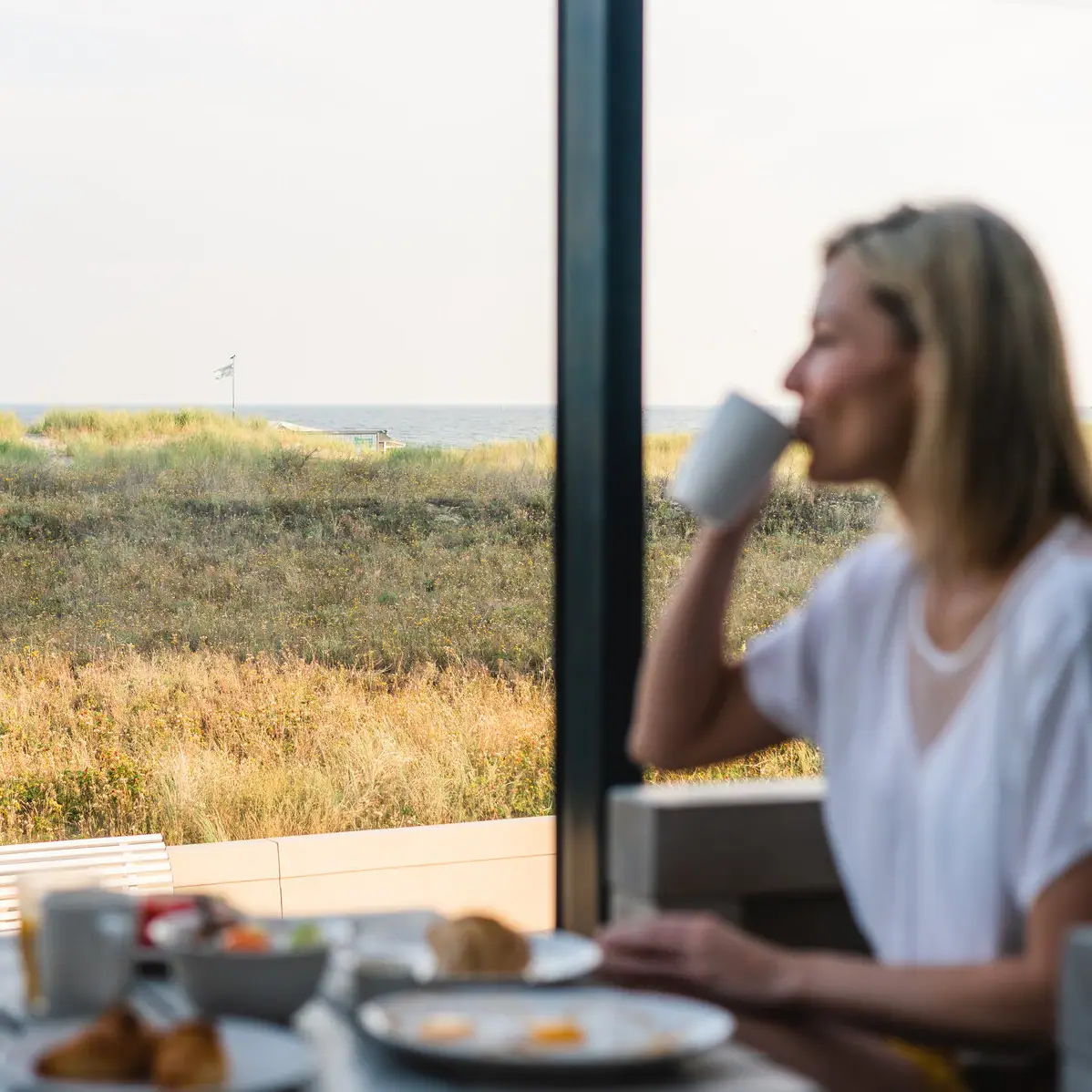 Frühstück mit Ostseeblick im aja Warnemünde Eine Frau sitzt an einem Tisch mit einer Tasse Kaffee.