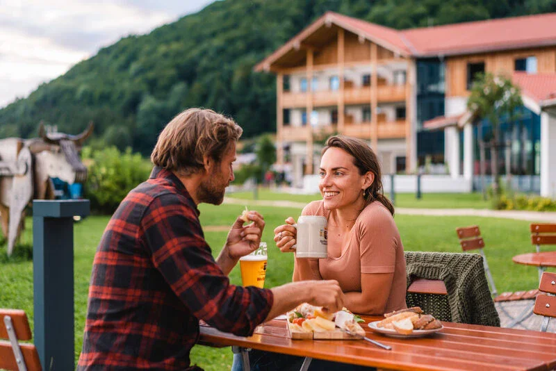 Ein Mann und eine Frau sitzen zusammen im Biergarten vom aja Ruhpolding, im Hintergrund ist das Hotel zusehen