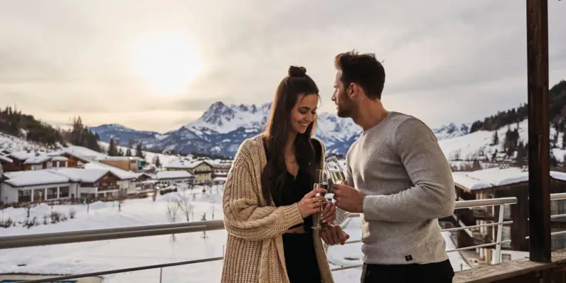 Ein Mann und eine Frau halten Weingläser auf einem Balkon mit schneebedeckten Bergen im Hintergrund.