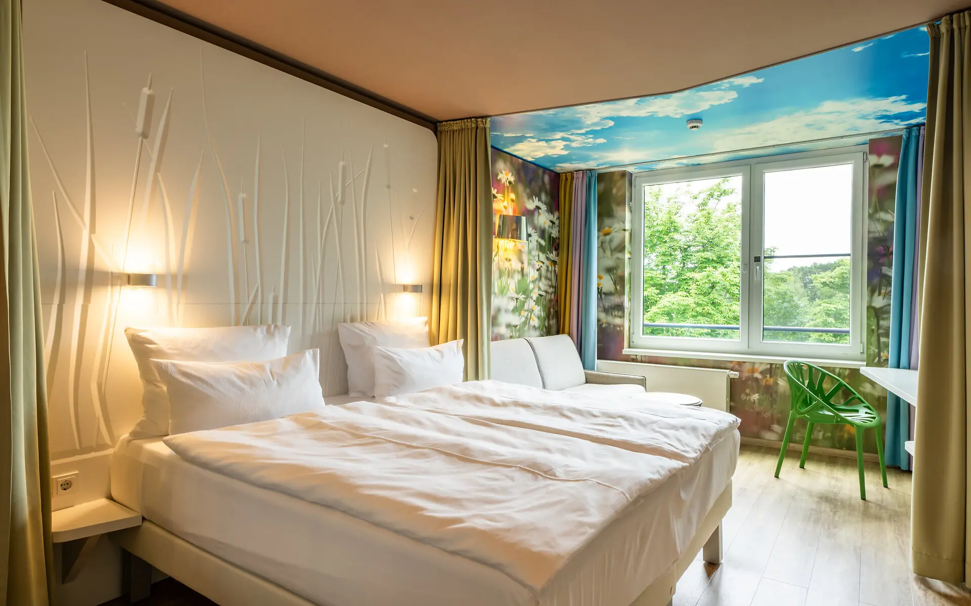Ein Hotelzimmer mit großem, weiß bezogenen Doppelbett, Sofa als Sitzmöglichkeit sowie einem Schreibtisch und großem Fenster mit Blick in die Natur. An der Wand beim Fenster ist die Tapete mit Blumen und Wolken bedeckt. 