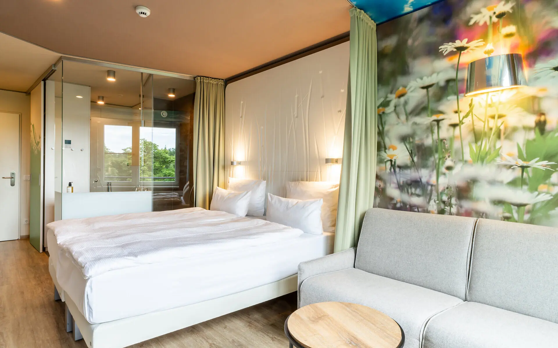 Ein Hotelzimmer in hellen Farben und naturbedruckter Wandtapete mit Doppelbett, Sofa und Tisch sowie teilweise Einsicht in das Badezimmer. 