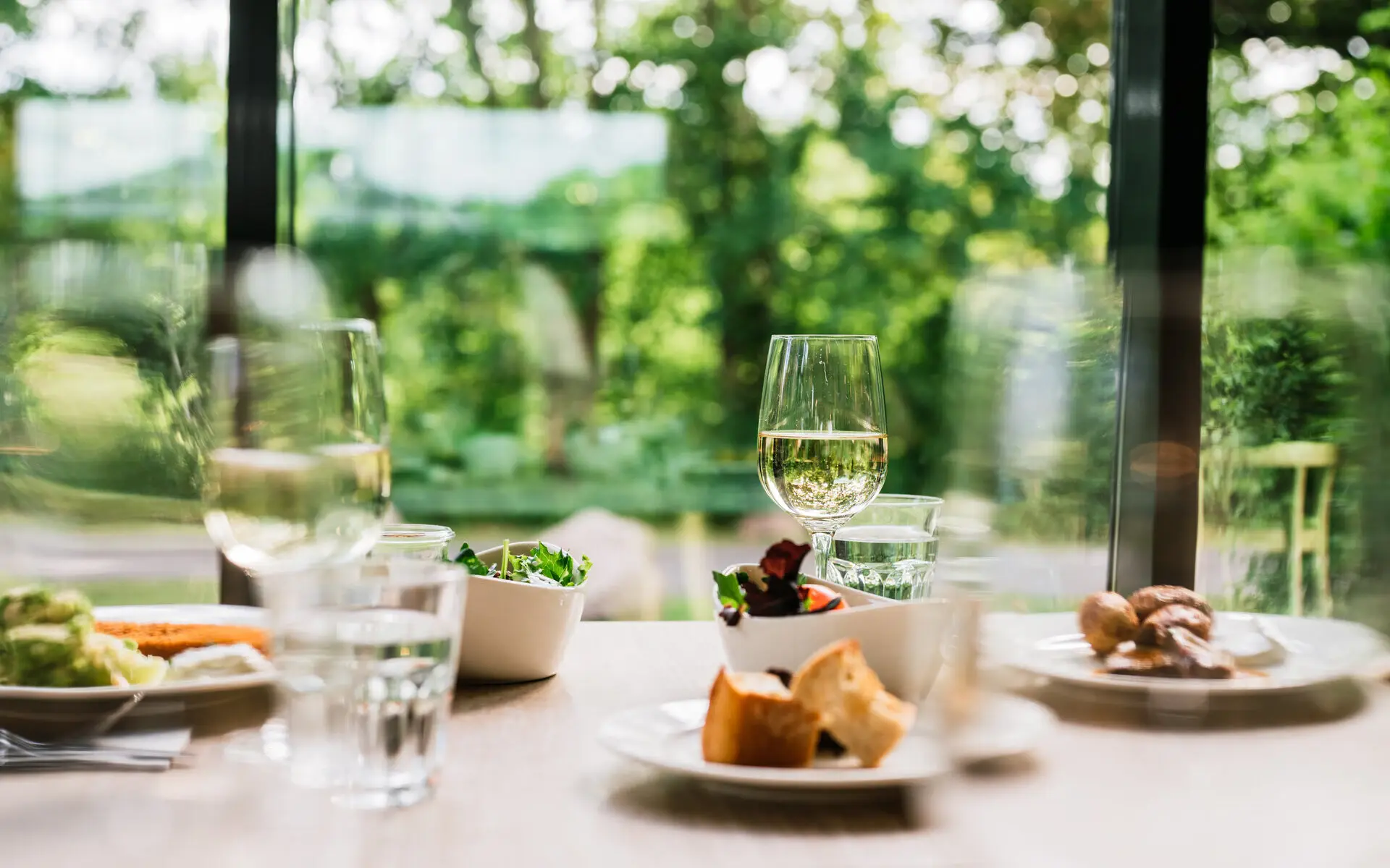 Table set with a variety of dishes and drinks, including wine glasses and plates, under a tree.