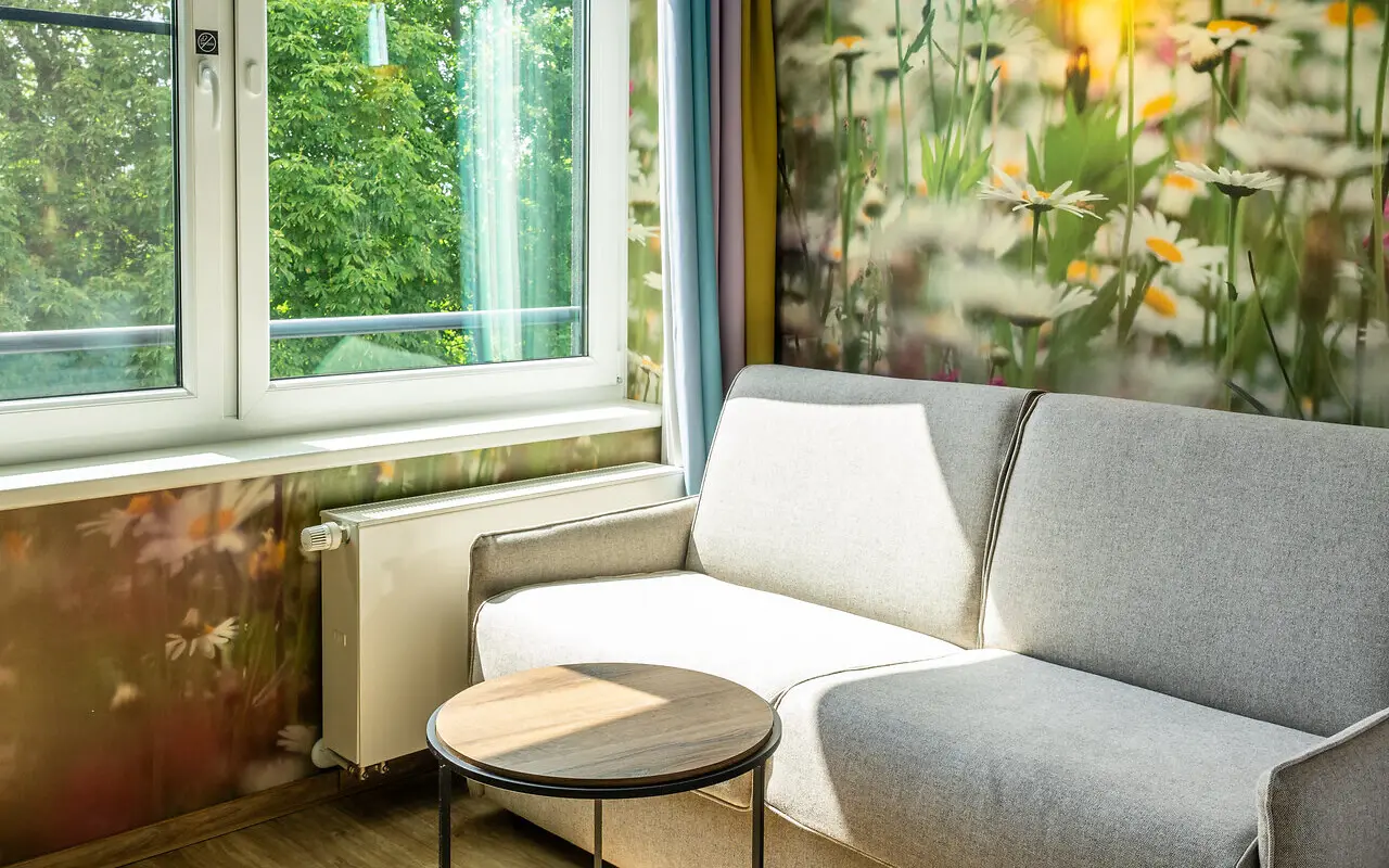 Ein gemütliches, hell graues zwei Personen Sofa vor einer naturgetreuen Blumentapete und neben einem großen Fenster, durch das warmes Sonnenlicht in das Hotelzimmer fällt und die Natur zu sehen ist. 