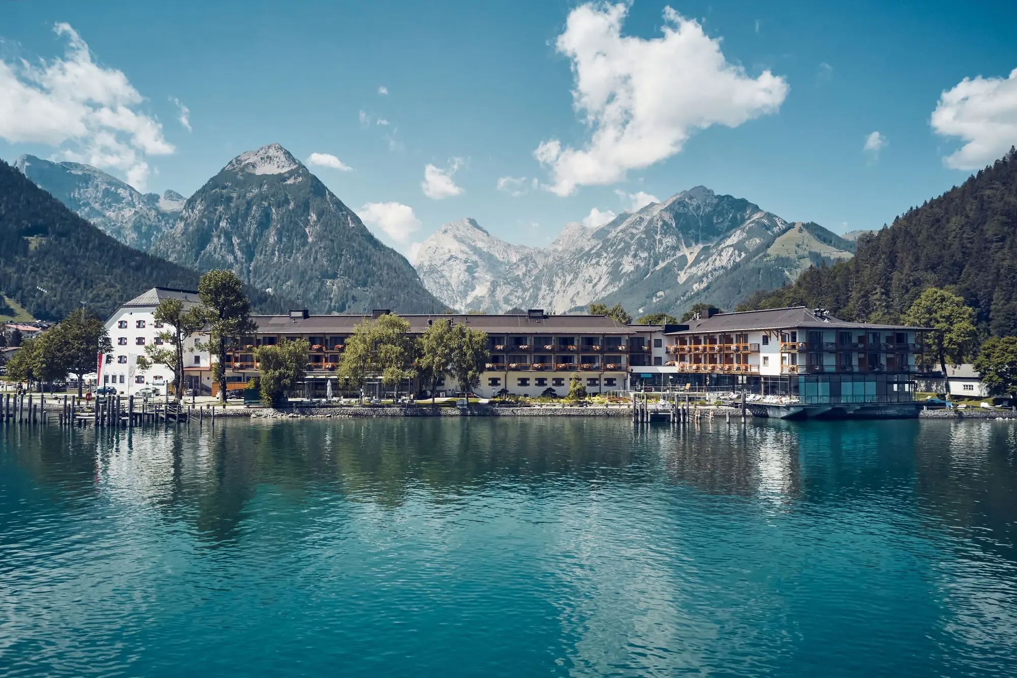 Blick auf das aja Achensee direkt am türkisblauen See, umgeben von imposanten Alpen und bewaldeten Hängen unter blauem Sommerhimmel.
